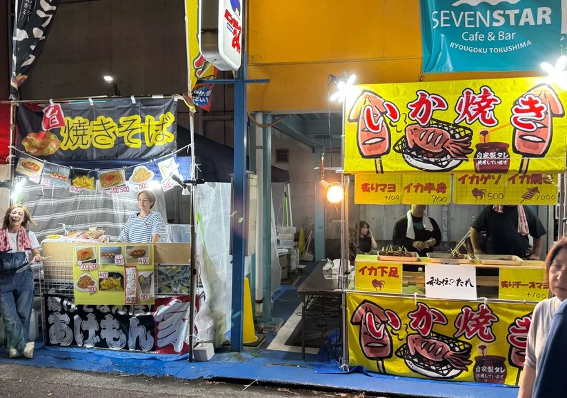 夜の駐車場に並んだ焼きそばといか焼きの屋台。黄色いのぼりが灯りに照らされている。