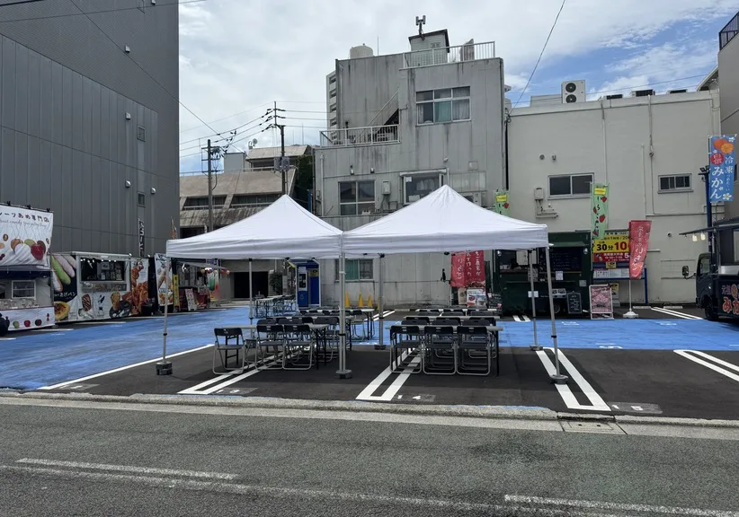 昼間の駐車場に白いテントを2張り設営し、長机とパイプ椅子を並べた様子。
